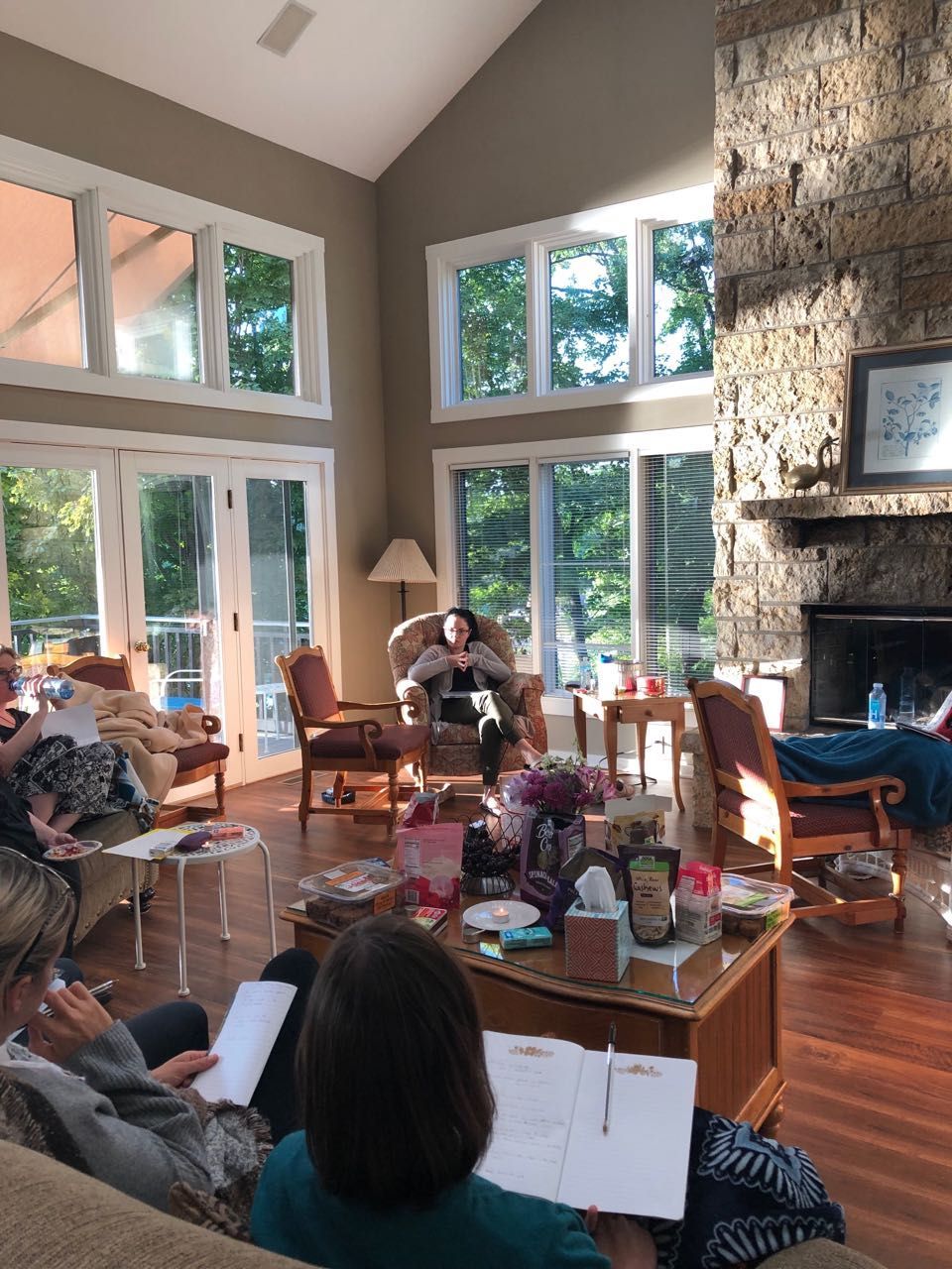 A group of people sits in a sunlit living room with a stone fireplace, holding notebooks while one person sits in a chair.