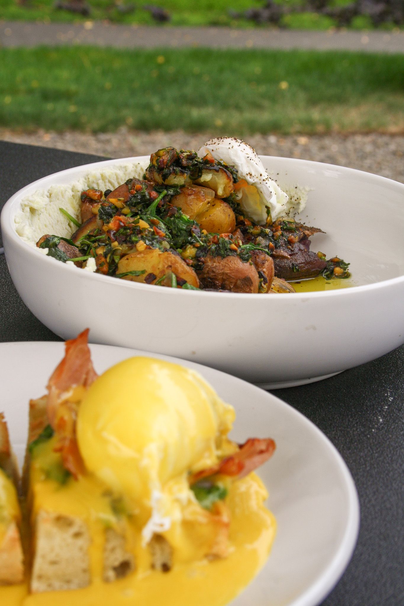 A bowl of sautéed vegetables and a plate of eggs benedict on toast sitting on a table outdoors.