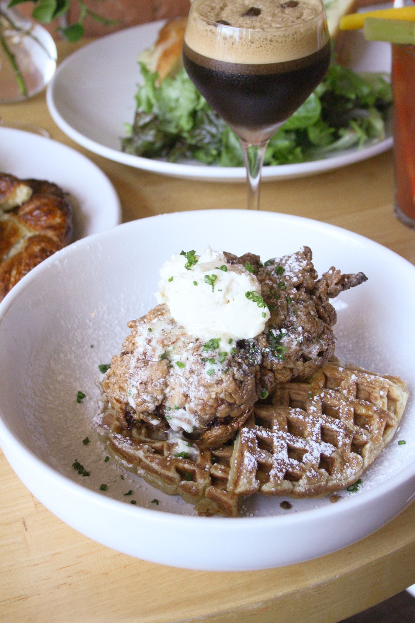 A bowl featuring fried chicken on a waffle topped with cream and herbs, with salads and a cocktail in the background.