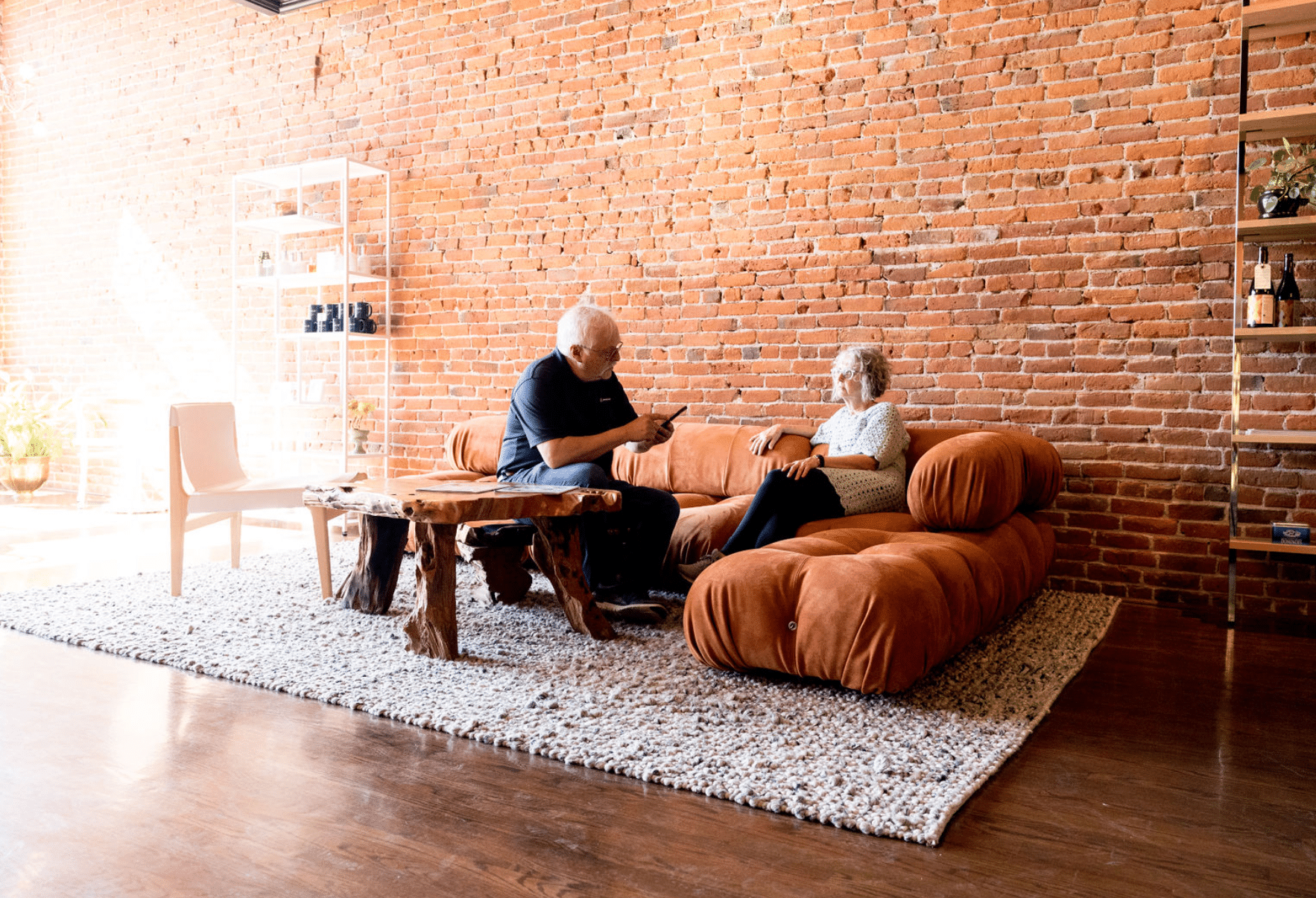 A man and a woman are sitting on a couch in a living room with a brick wall.