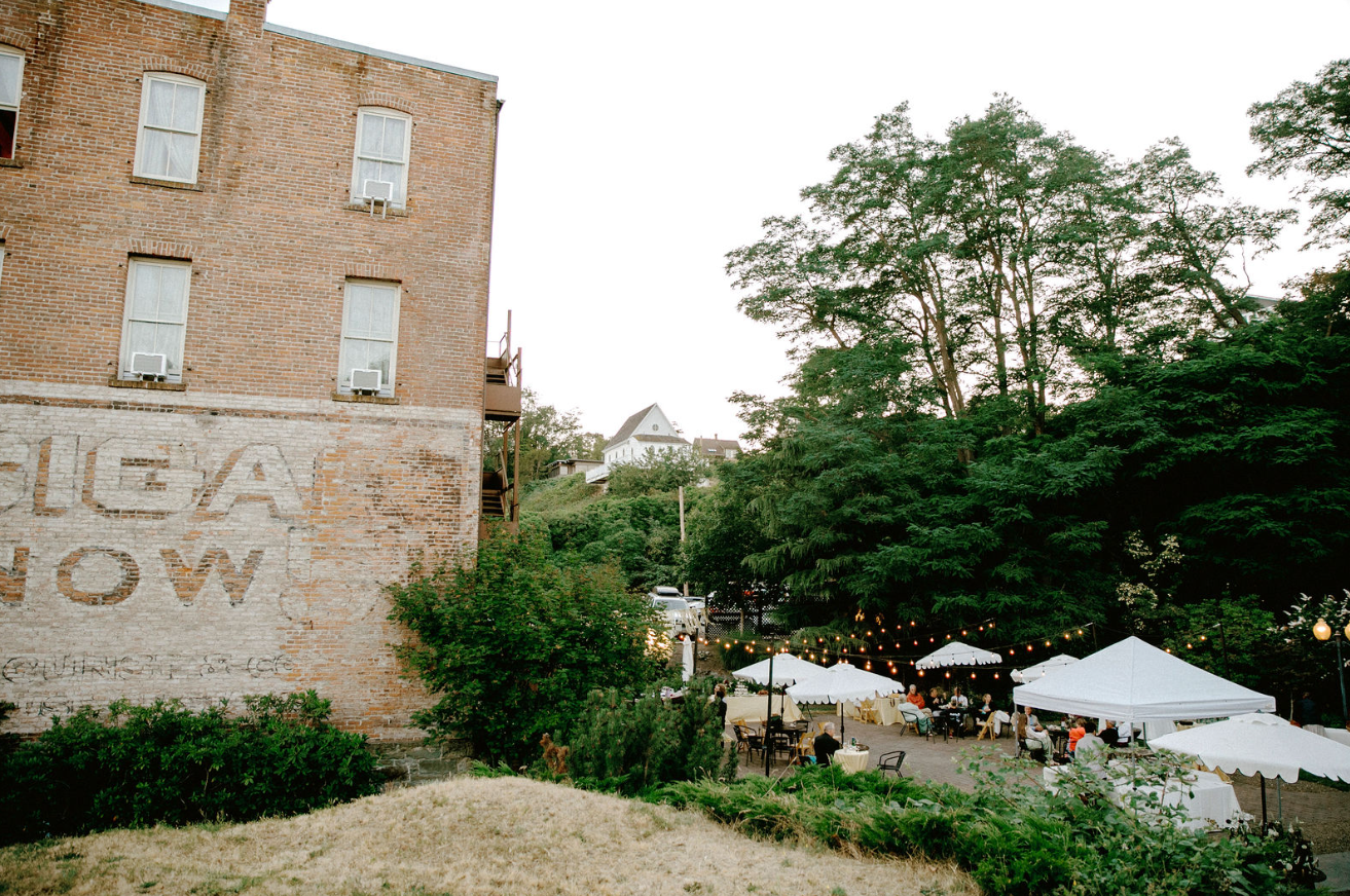 A weathered brick building with faded text next to a tree-lined outdoor space featuring white event tents and guests.