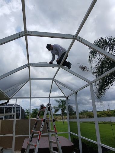 Man works on a white screened enclosure roof; ladder nearby; cloudy sky, green yard in background.