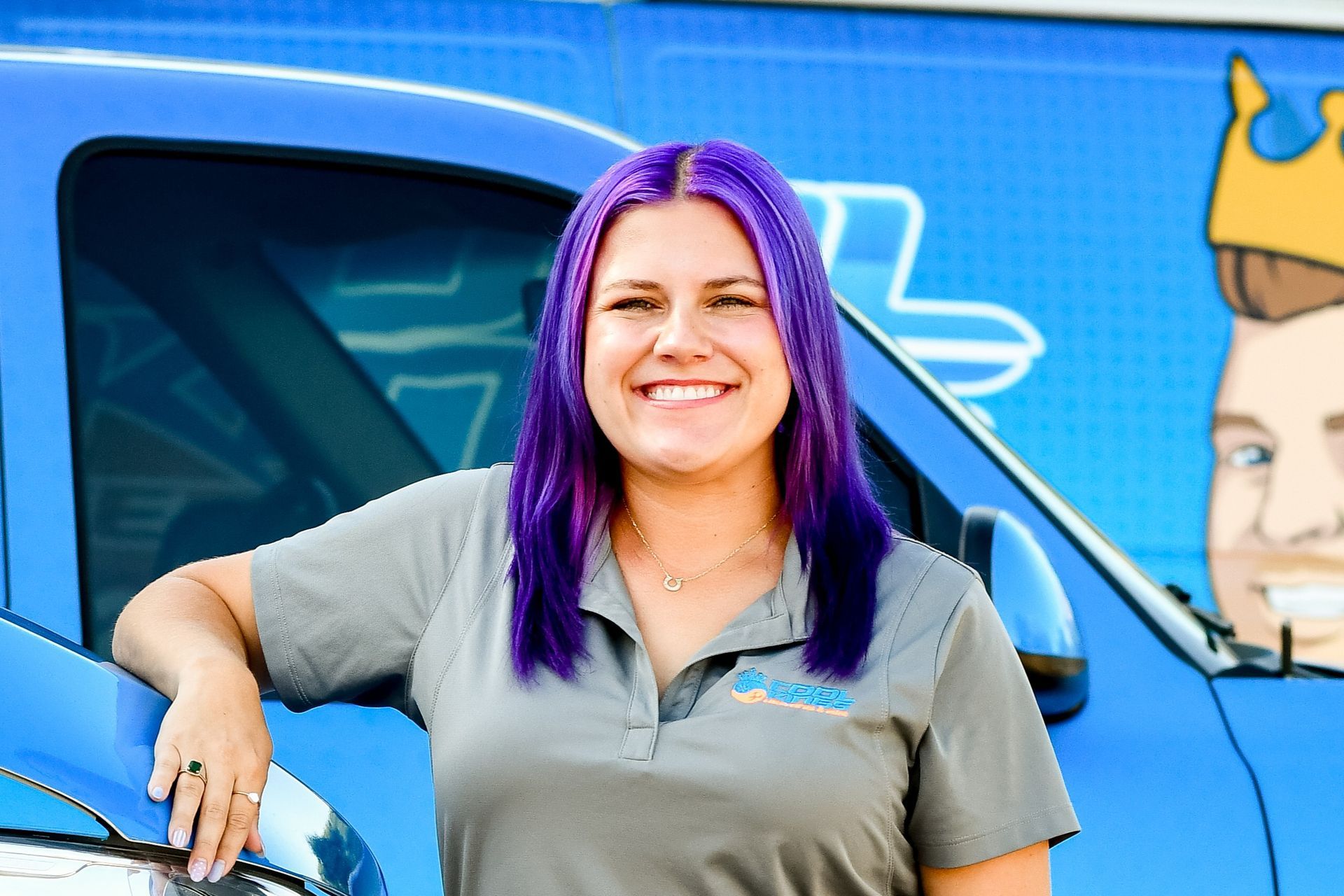 Smiling person with purple hair leaning on a blue van with a cartoon mural