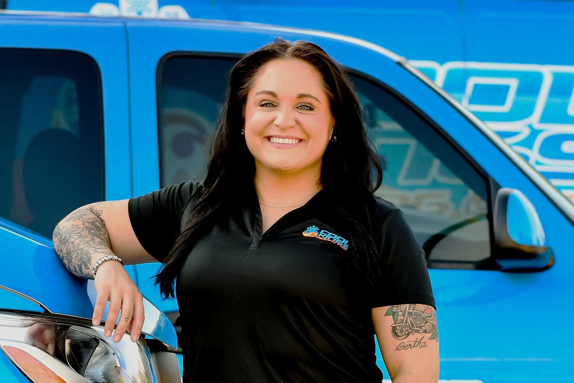 Woman smiling beside a bright blue van, wearing a black shirt with visible tattoos on her arms.