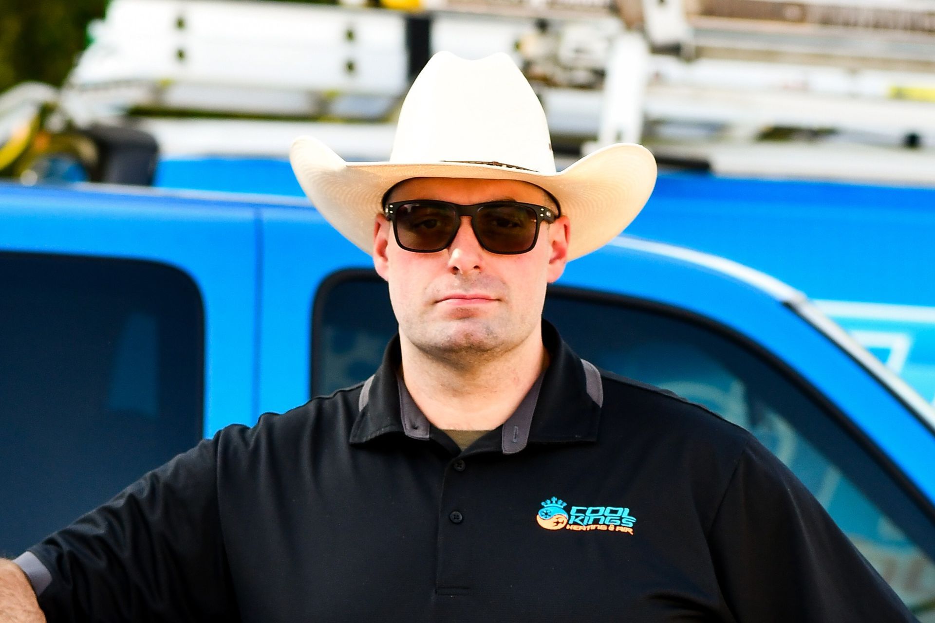 Man in white cowboy hat and sunglasses standing before a blue truck, wearing a black shirt.