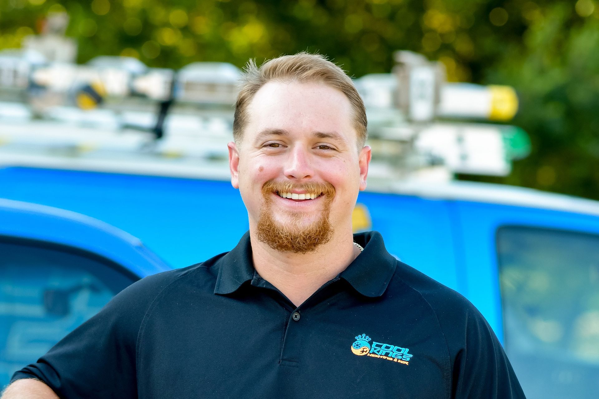 Smiling man in dark polo standing beside a blue vehicle outdoors