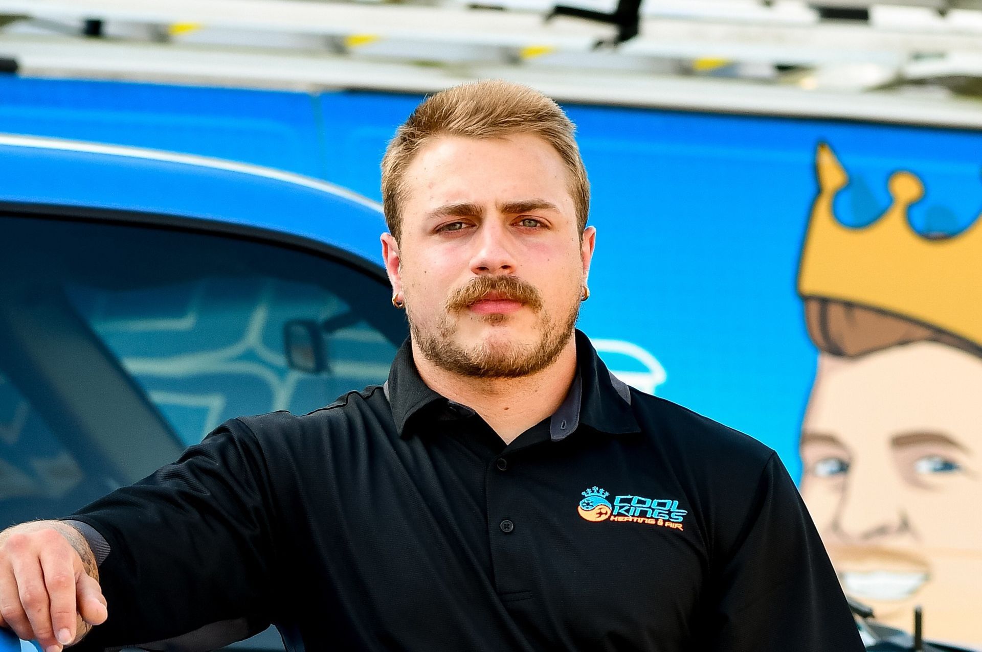 Man standing beside a blue car with a cartoon crown logo, wearing a black shirt, in front of a colorful mural