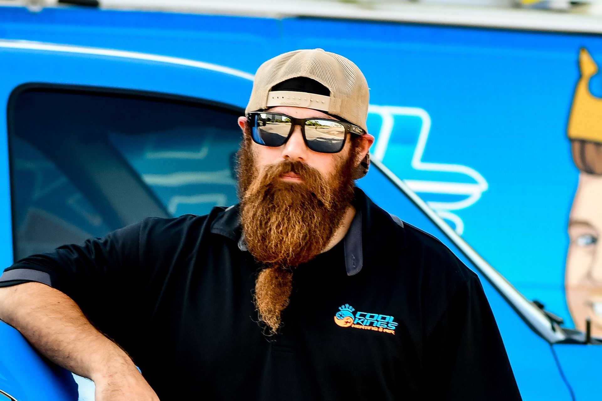 Bearded man in sunglasses and cap leaning on a blue truck with a logo on his shirt