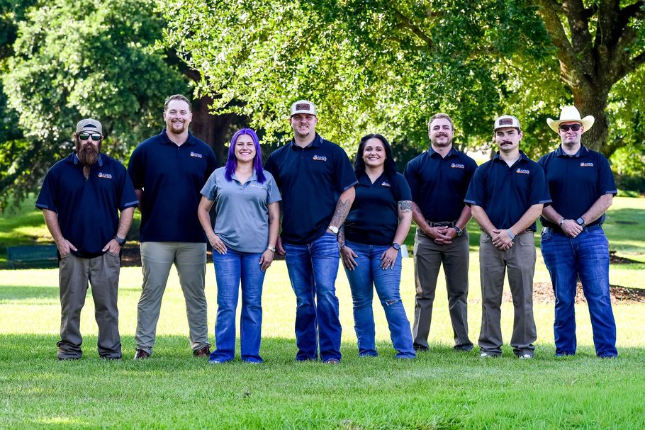 Group of seven people standing outdoors on grass, wearing navy shirts in front of leafy trees.