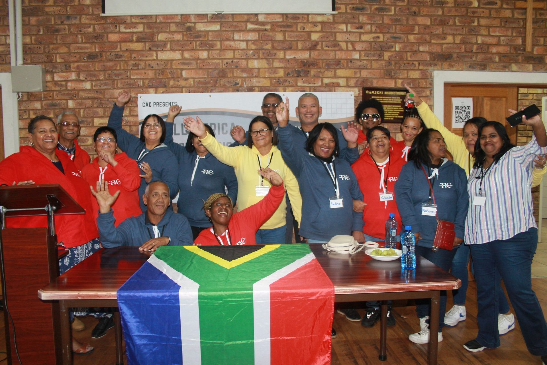 Group of people celebrating, South African flag on table, brick wall background.