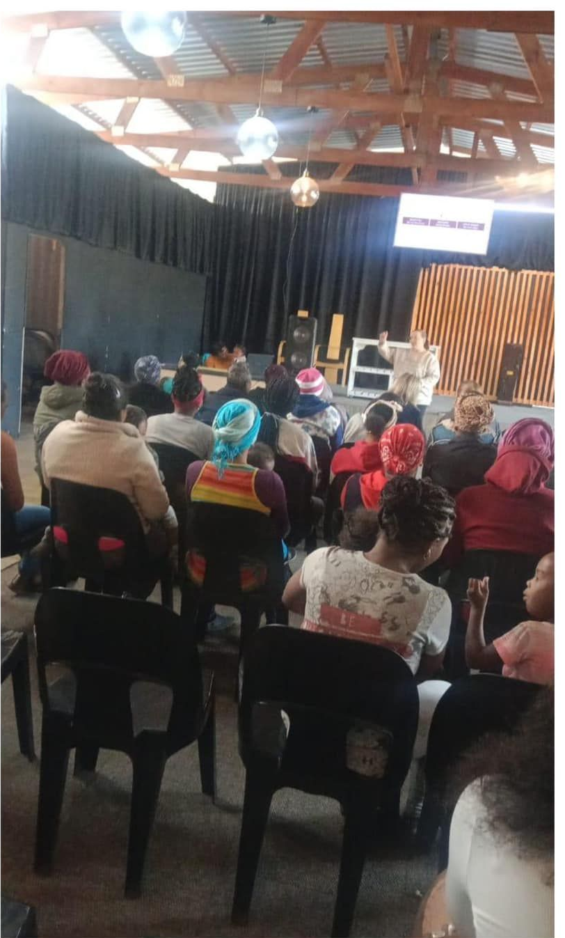 Audience seated in a community hall, watching a presentation on a screen.