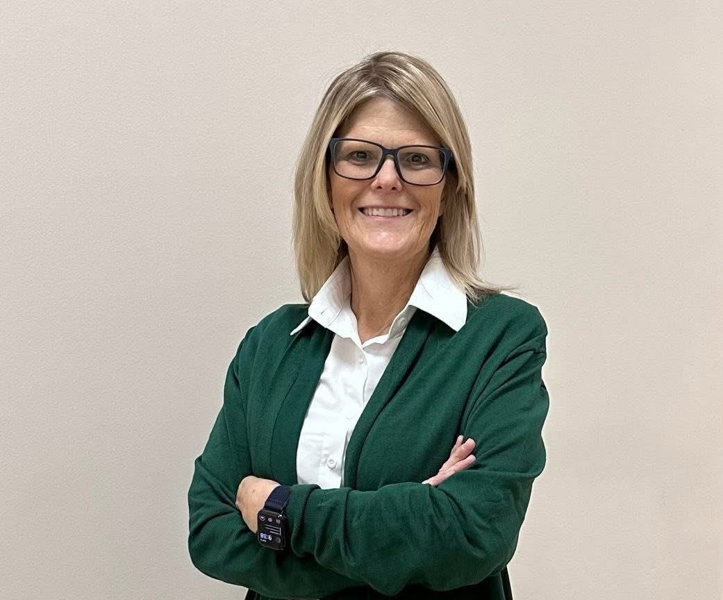 Woman with glasses, arms crossed, wearing a green sweater and white shirt, smiling.