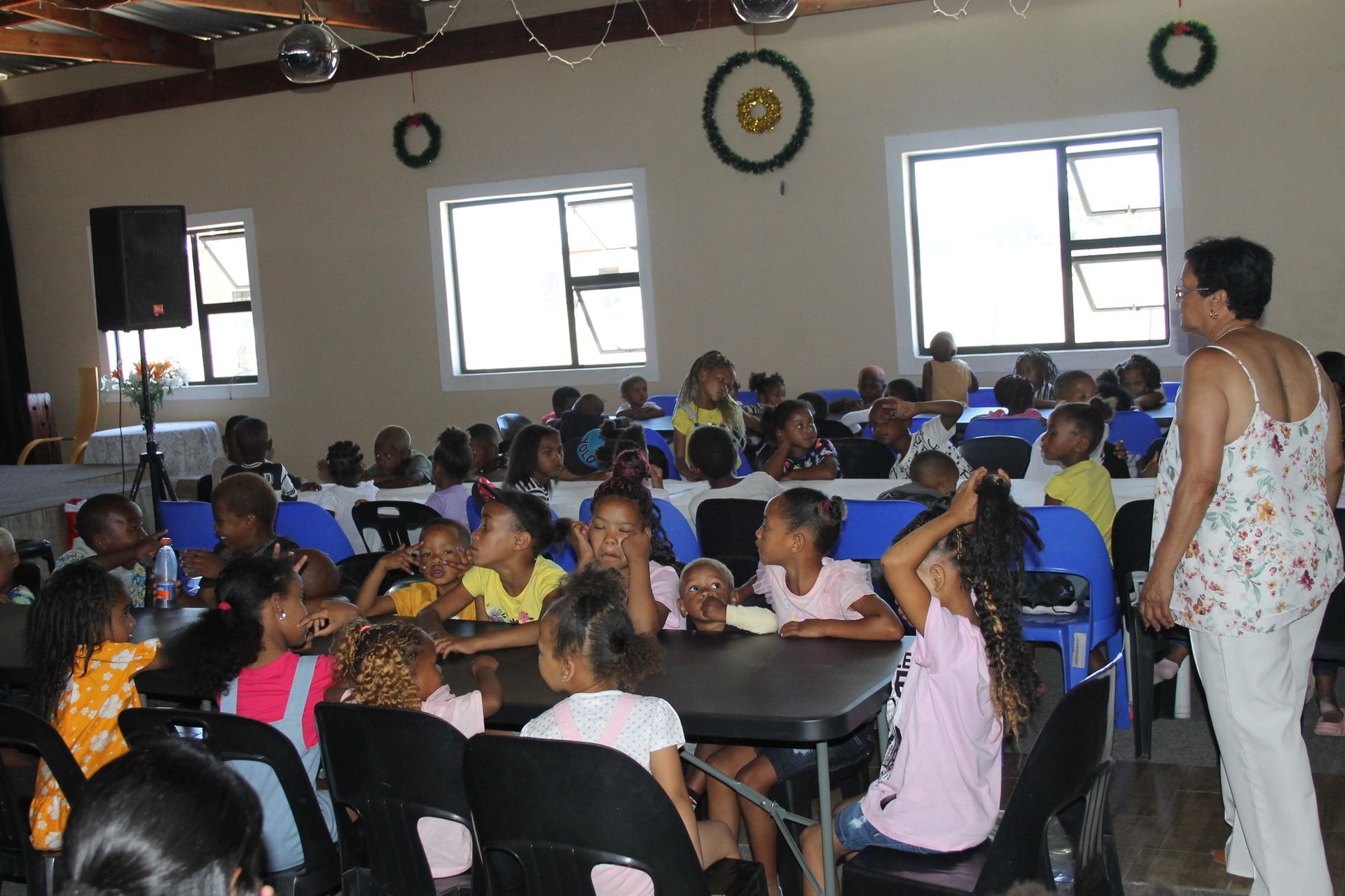 Children seated at tables, listening to a woman in a room with windows.
