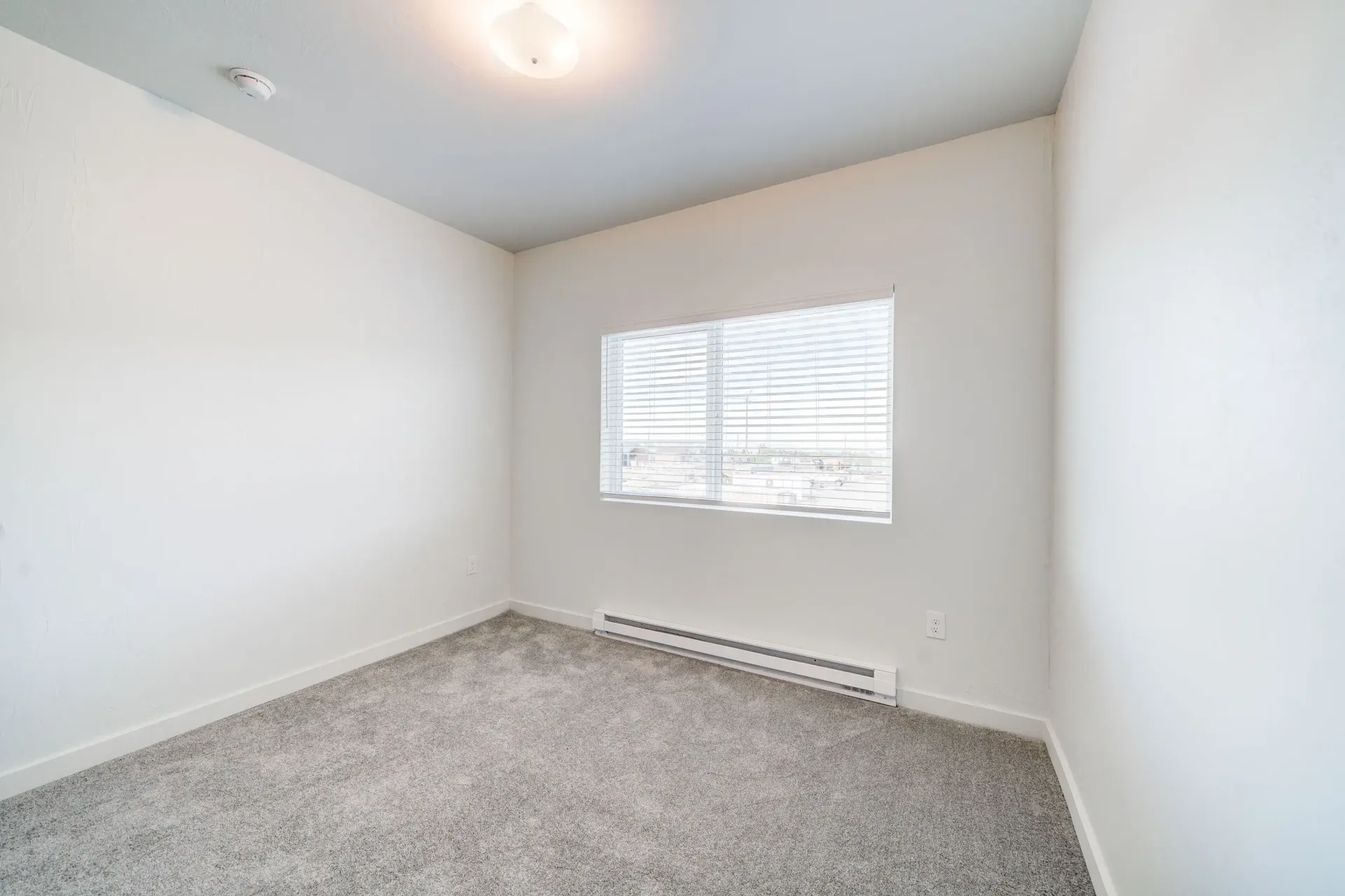 Empty bedroom with white walls, a window with blinds, carpet, and a baseboard heater.
