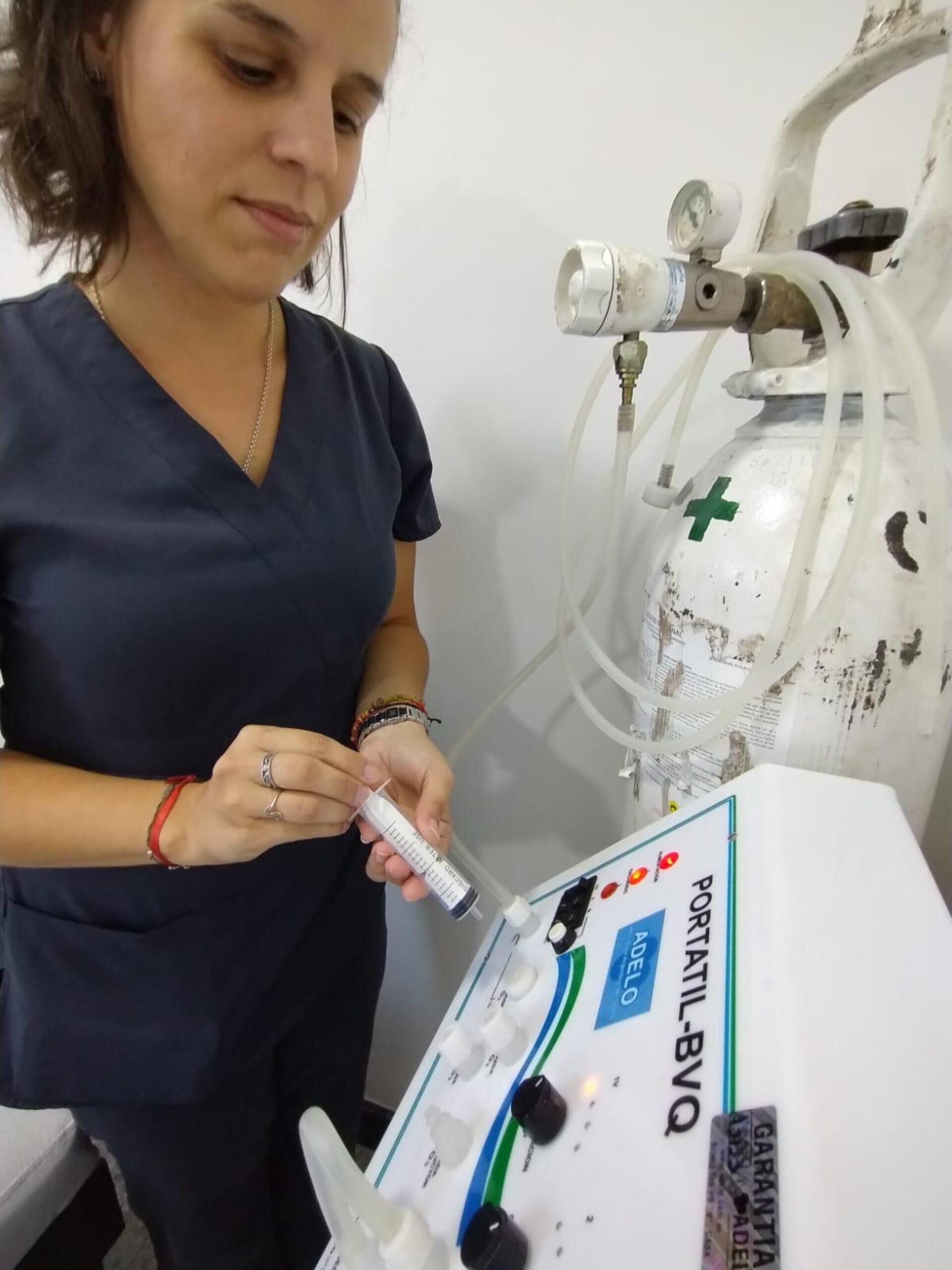 Un profesional sanitario con bata azul prepara una jeringa en una máquina conectada a un tanque de oxígeno.