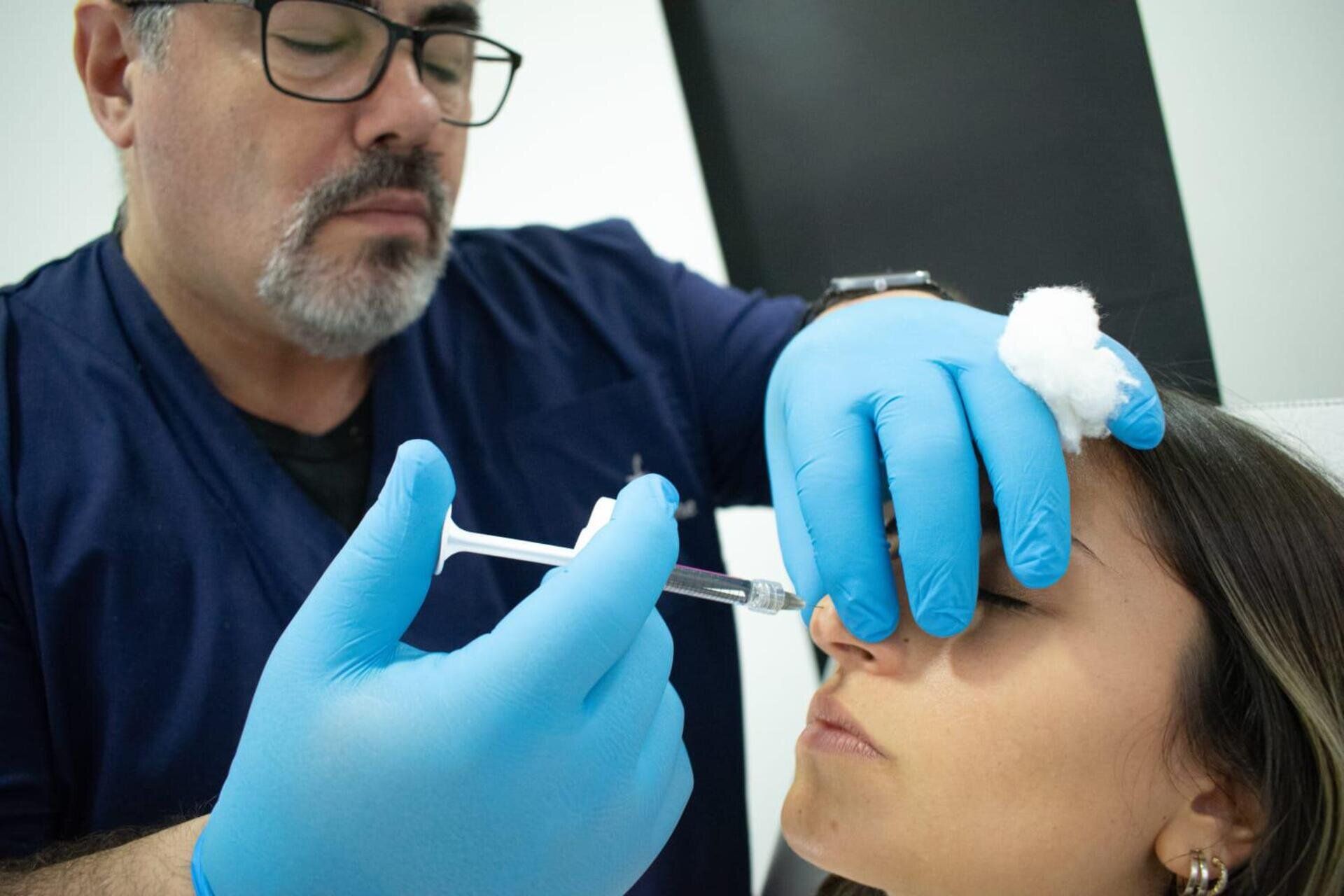 Un profesional con bata y guantes azules realiza un procedimiento de relleno nasal no quirúrgico con una jeringa.