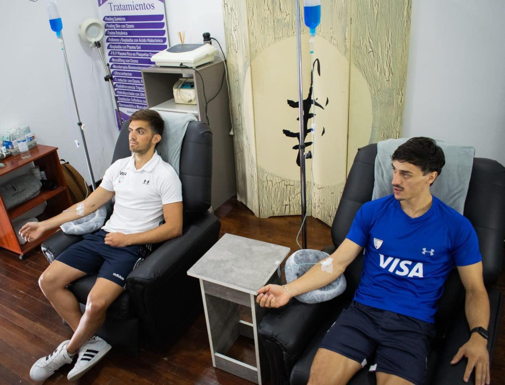 Dos personas están sentadas en sillas recibiendo terapia intravenosa