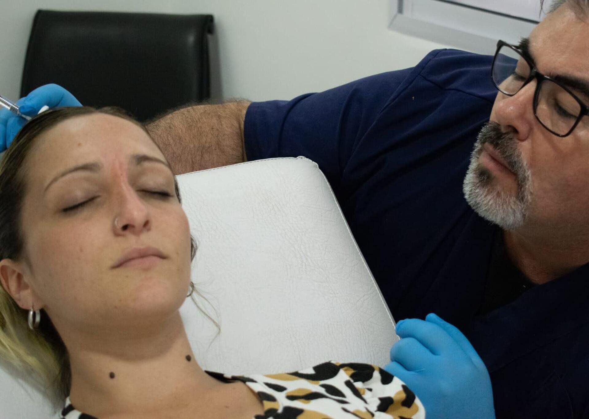 Un profesional con bata y guantes azules realiza una inyección cosmética en la frente de un paciente en una clínica.