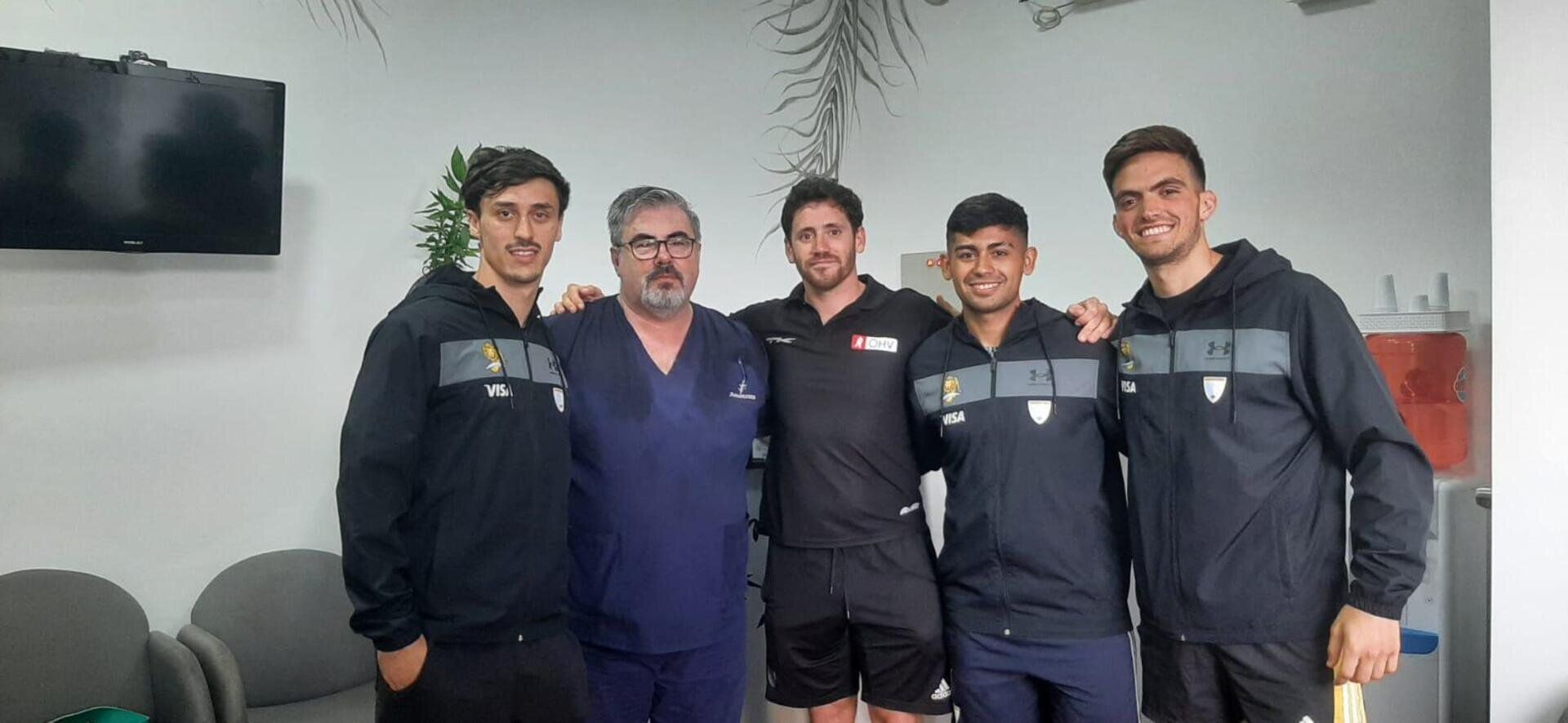 Cinco personas están de pie en una habitación, cuatro con chaquetas deportivas oscuras iguales y una con uniforme médico azul