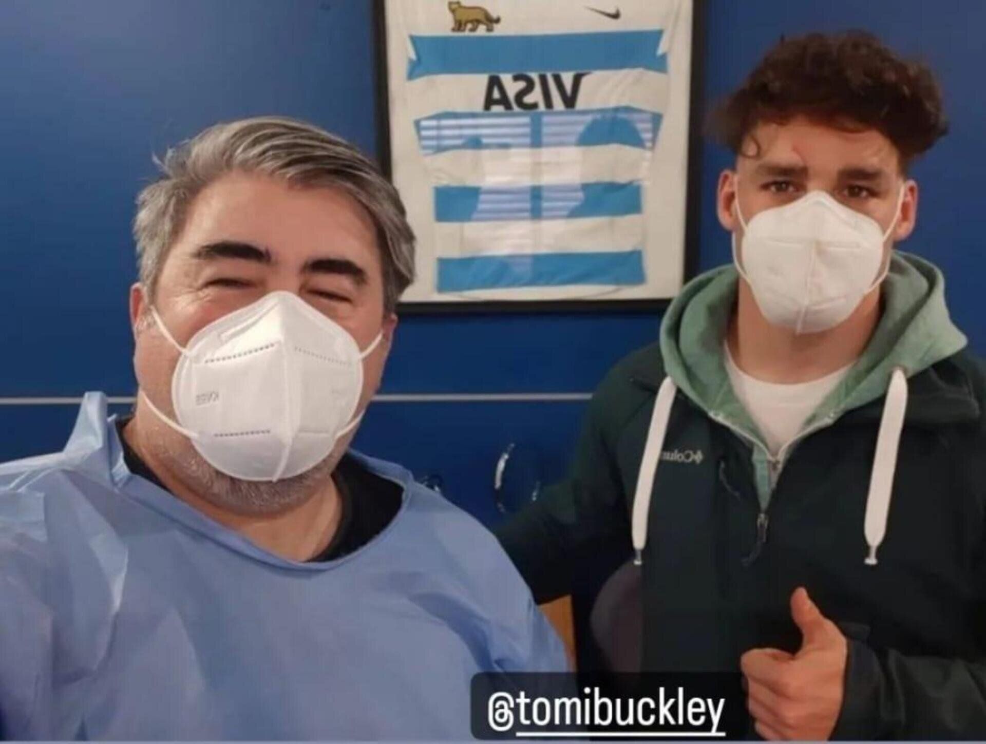 Dos personas con mascarillas posan para una selfie frente a una camiseta deportiva azul y blanca enmarcada.