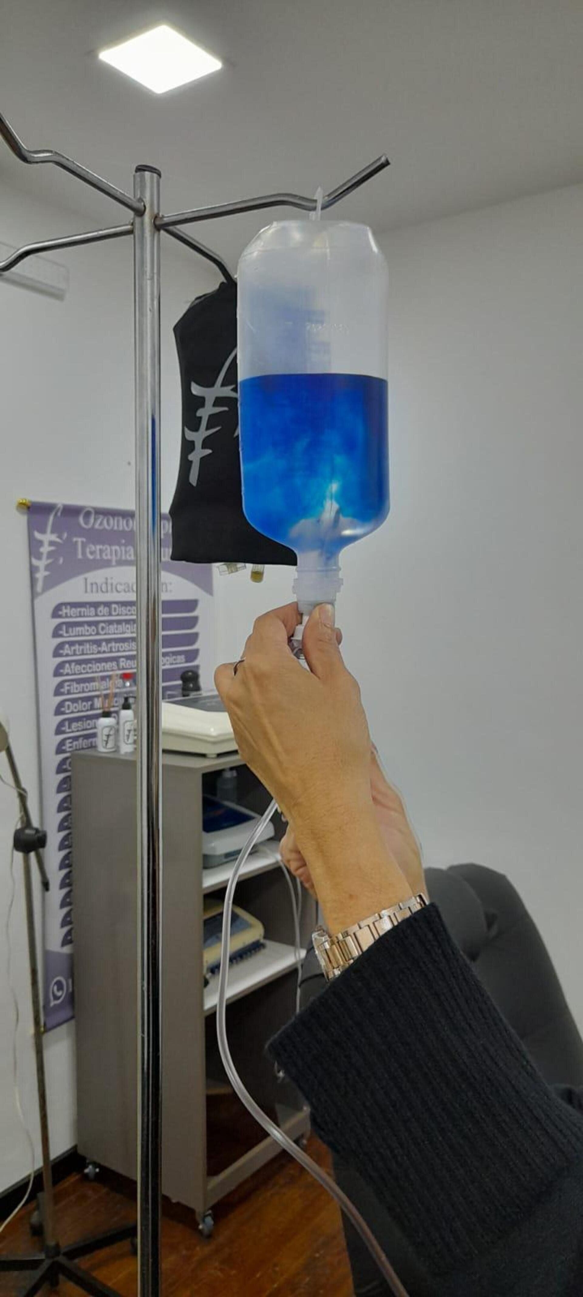 En una habitación, una persona sostiene en su mano una bolsa de suero llena de líquido azul que cuelga de un soporte médico.