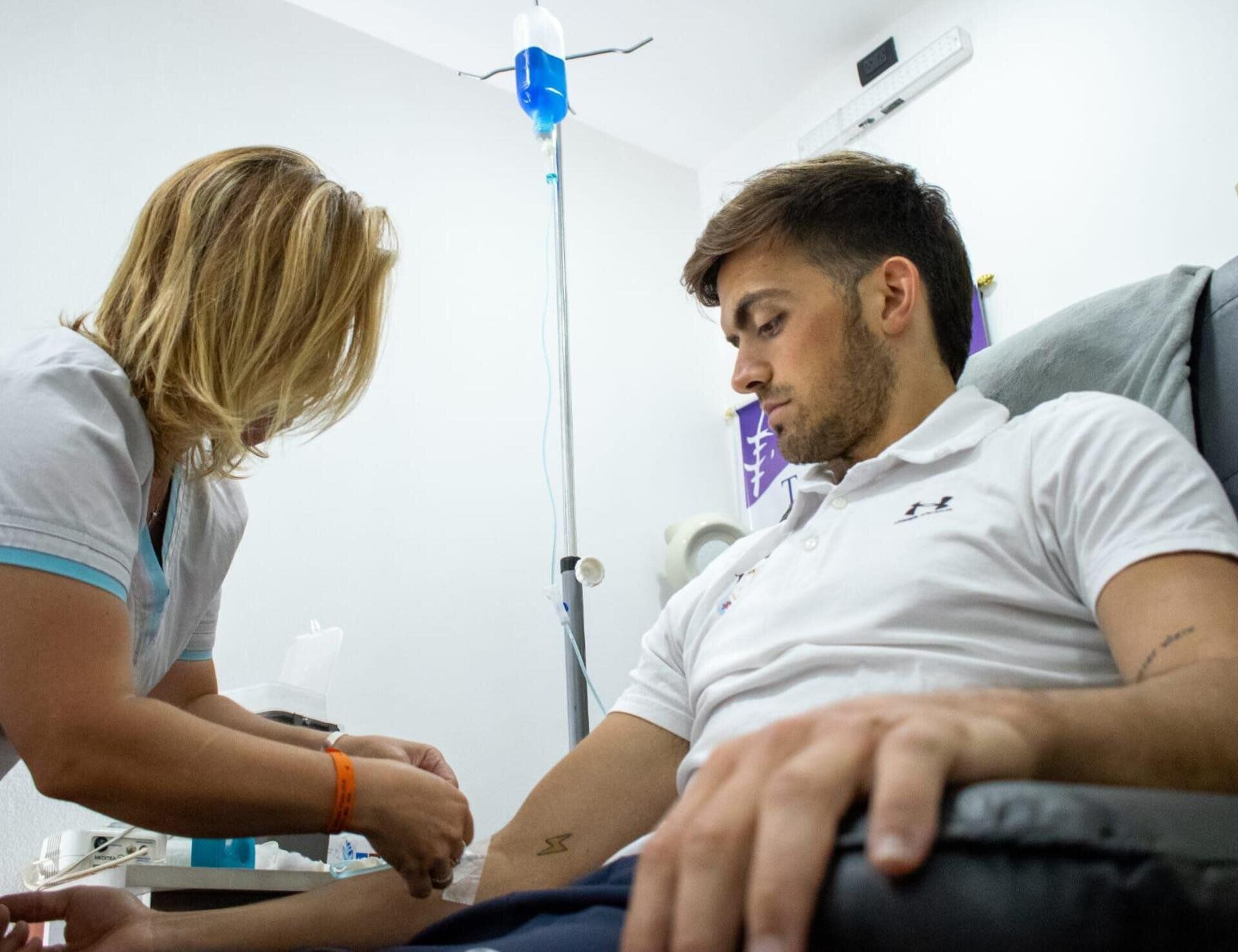 Un profesional sanitario con uniforme blanco prepara una vía intravenosa para un paciente sentado en una silla clínica.