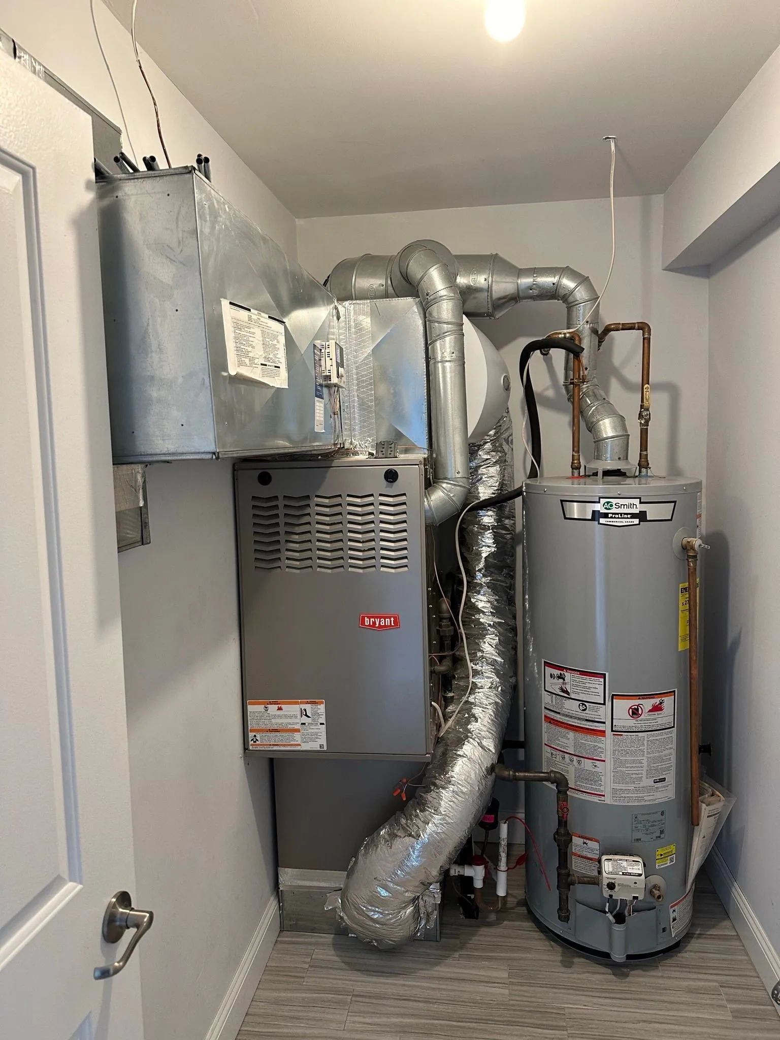 A small utility room with a furnace, water heater, and ductwork. Silver and gray appliances against light gray walls.
