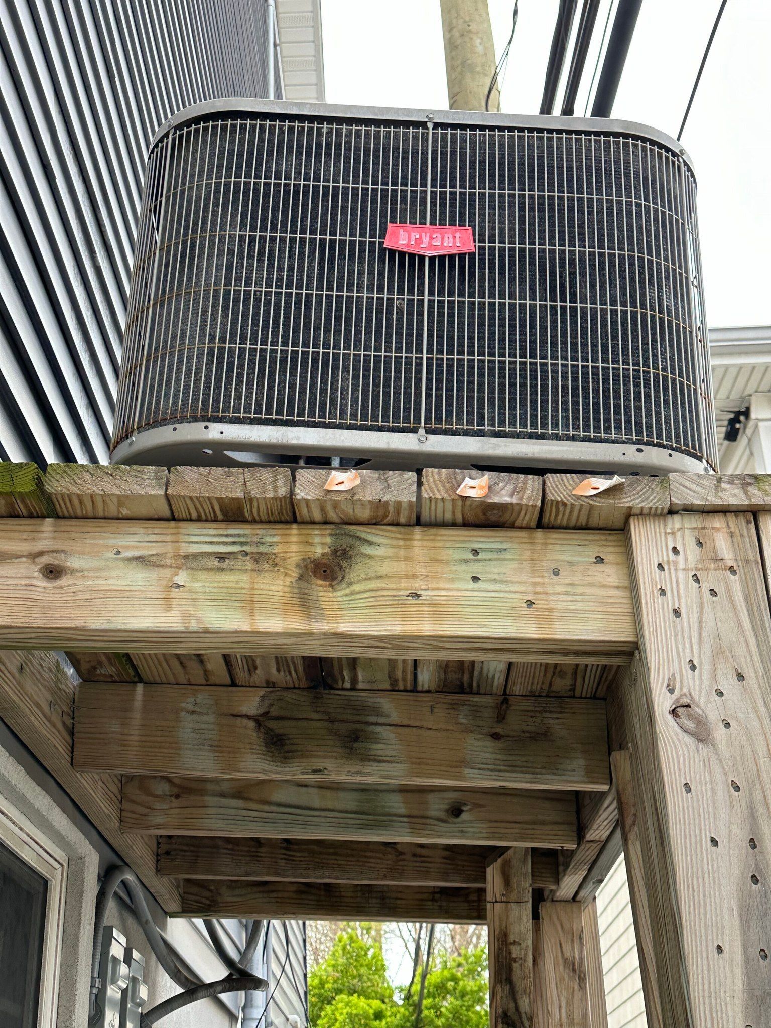 Air conditioning unit on a weathered wooden deck, against a building with siding and overhead power lines.