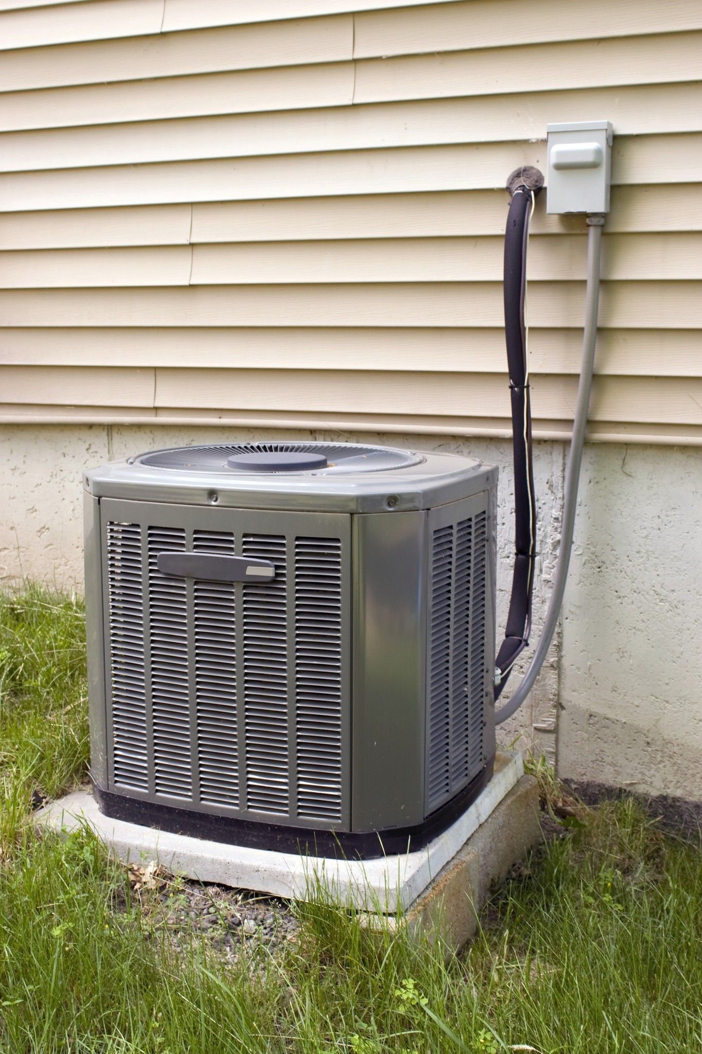 Central air conditioning unit outside a beige house with beige siding, gray unit, and conduit.