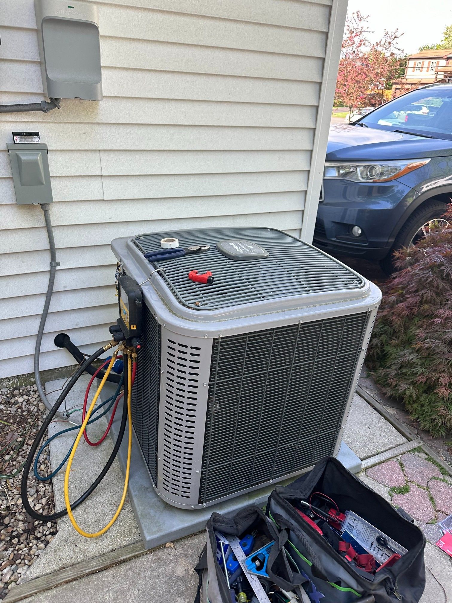 HVAC unit outside a house, being serviced with gauges attached. A gray car is in the background.