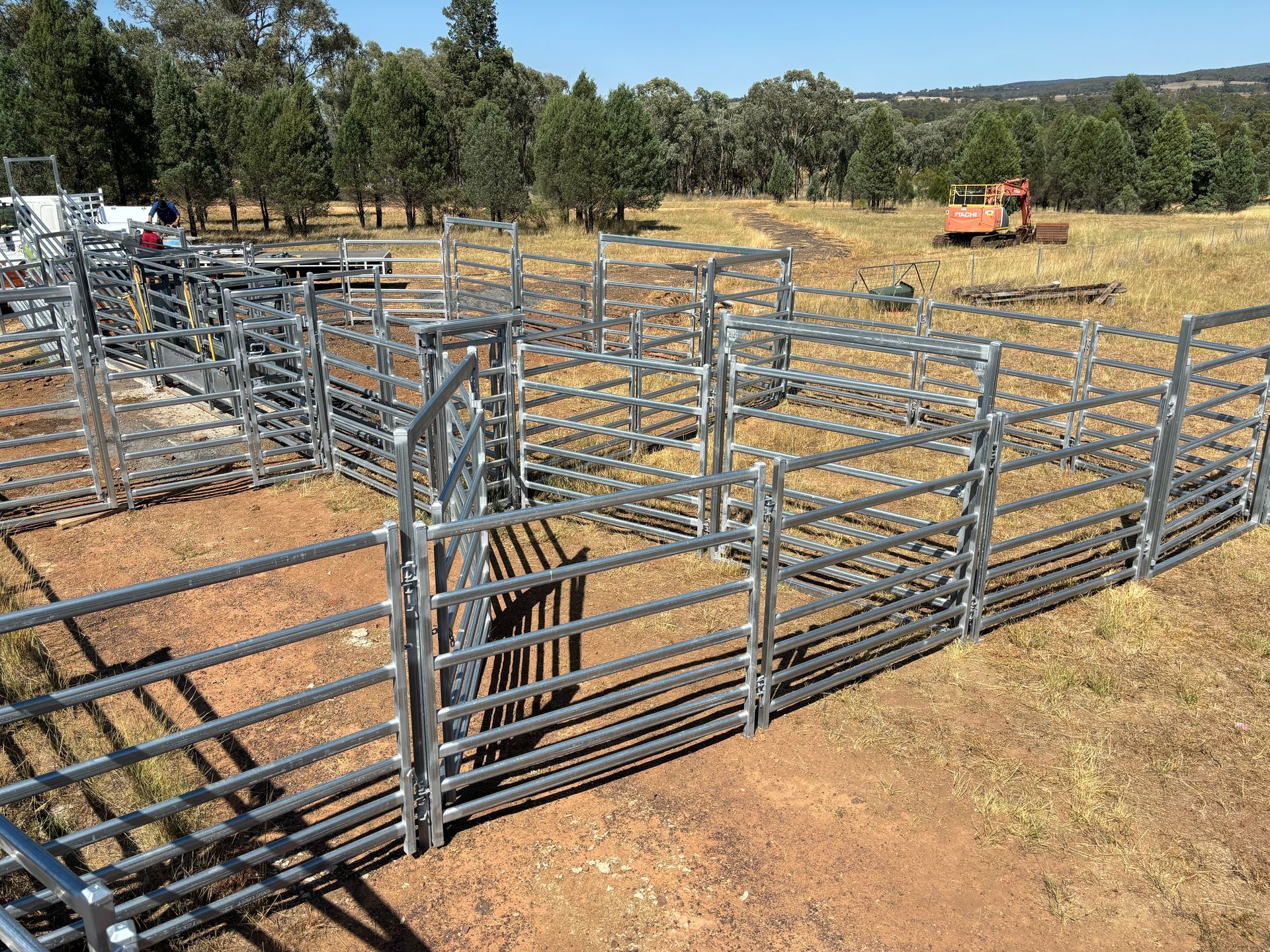 Metal livestock yard with gates, set in a field with trees and machinery in the background.