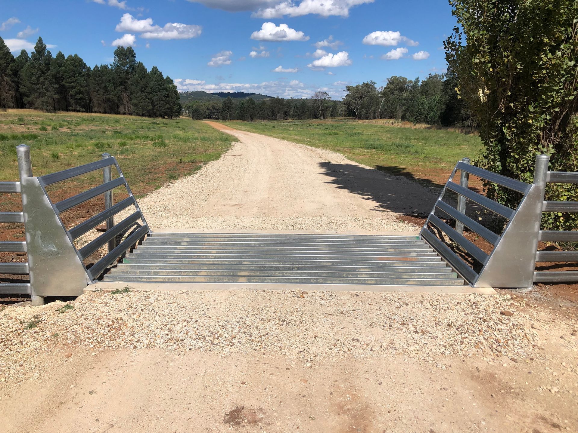 Metal cattle grid on a gravel road, leading to a field with trees in the background under a blue sky.