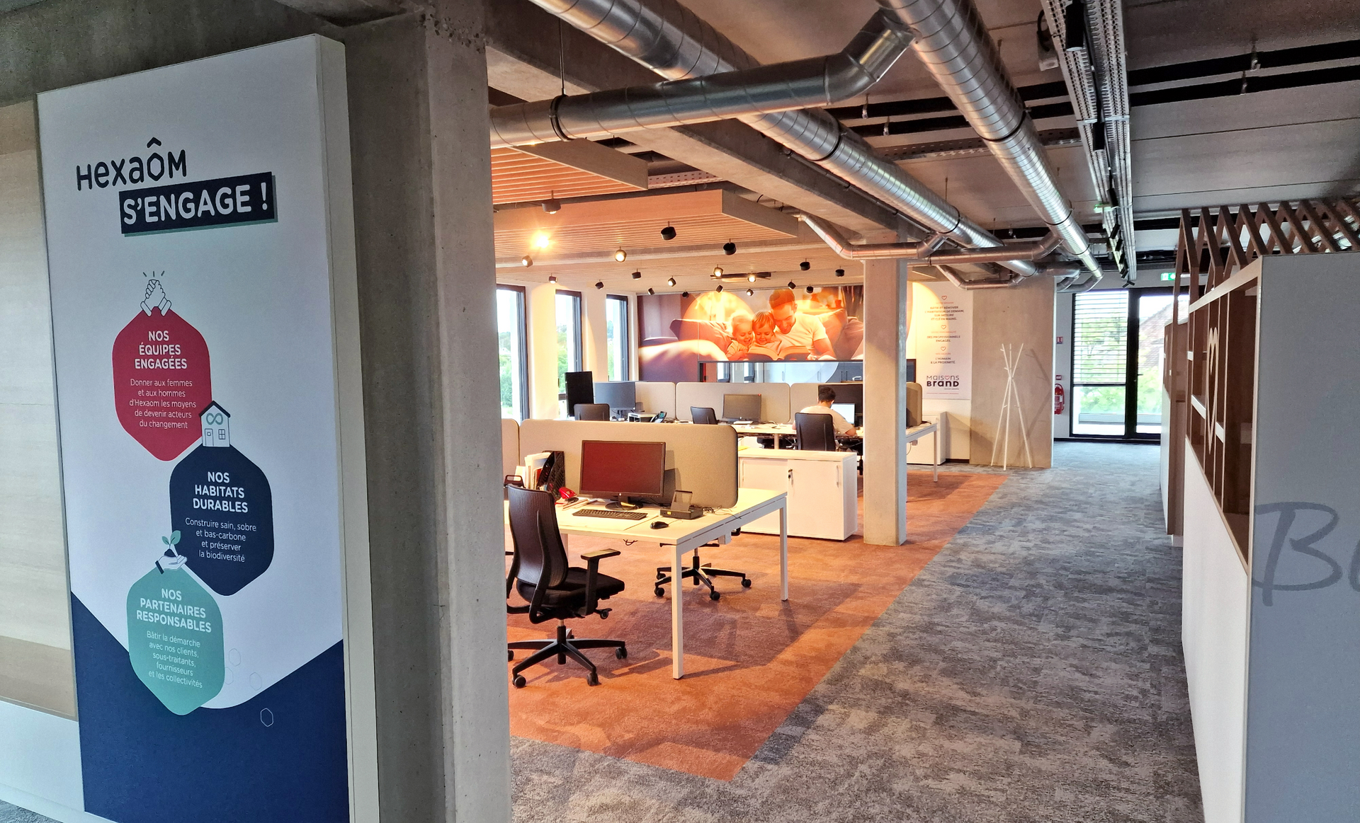 Office interior with desks, chairs, and colorful banner. Sunlight and orange carpet. Concrete columns and exposed pipes.