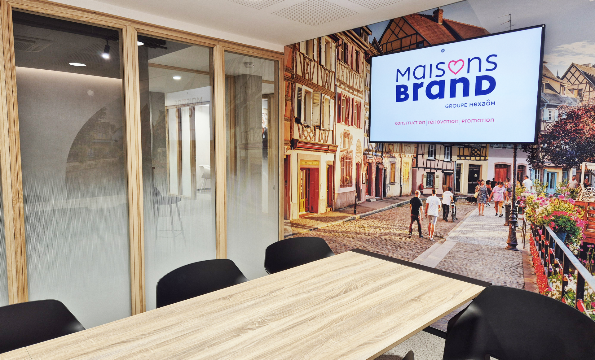 Meeting room with wood table, black chairs, and a screen displaying a company logo. Wall mural depicts a town square.