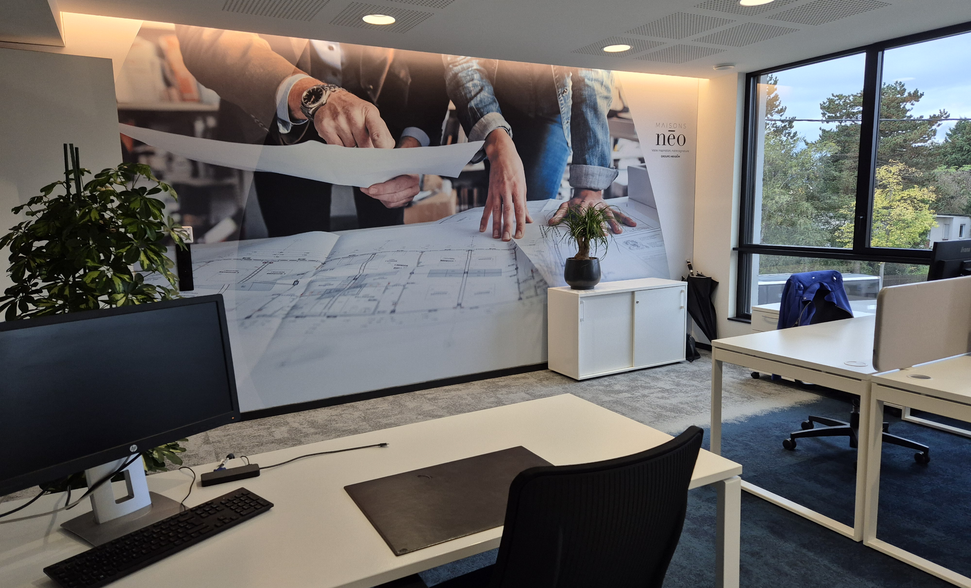 Modern office interior with desks, a large mural of a person working on blueprints, and a window overlooking greenery.