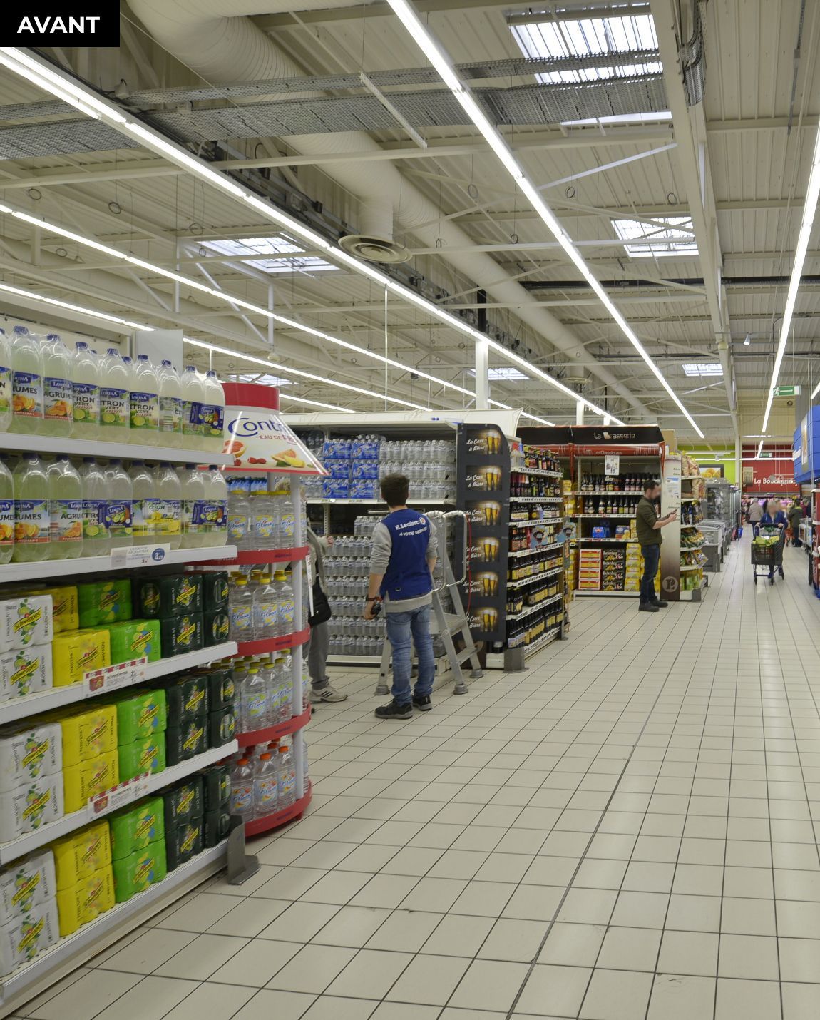 AVANT TRAVAUX Une allée de supermarché avec des étagères remplies de produits et un client debout près du centre.