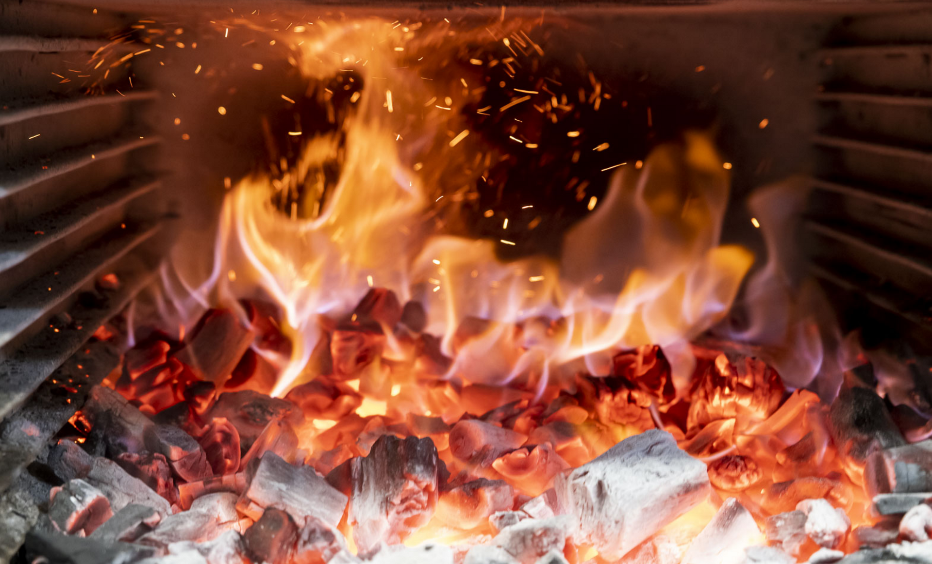 Burning charcoal in a furnace; orange flames and sparks rise from the red-hot embers.