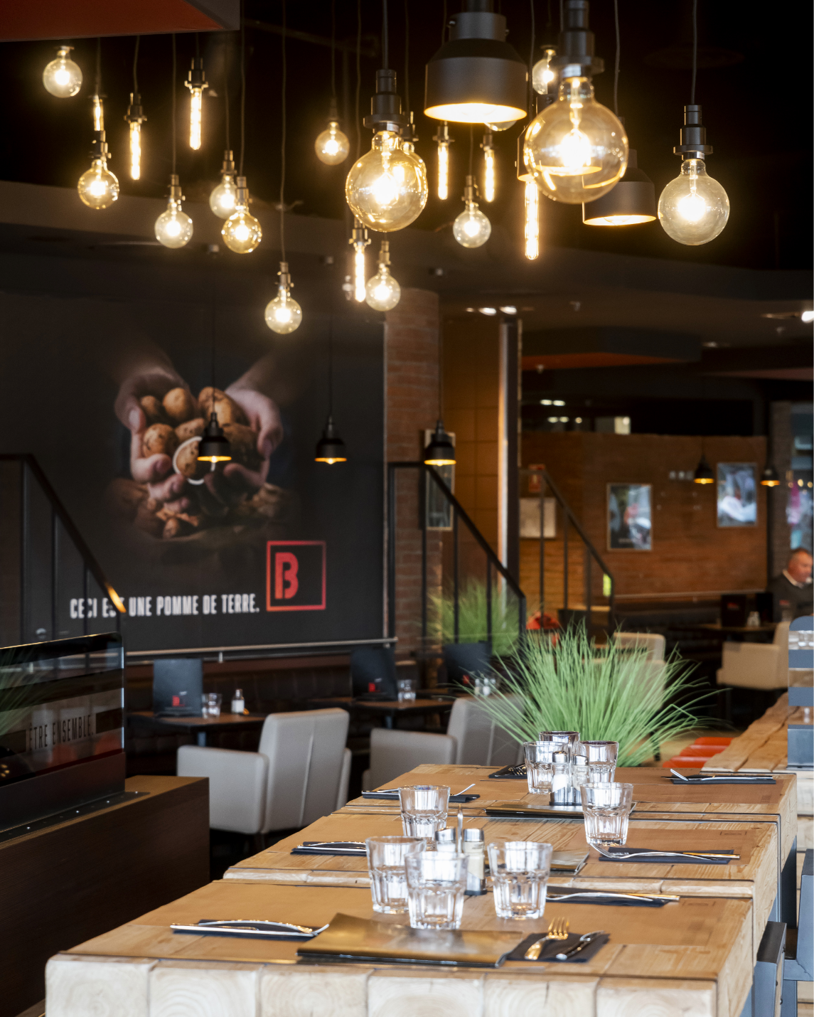Restaurant interior: wooden tables set with glasses, overhead Edison bulbs, brick wall with a mural and a staircase.