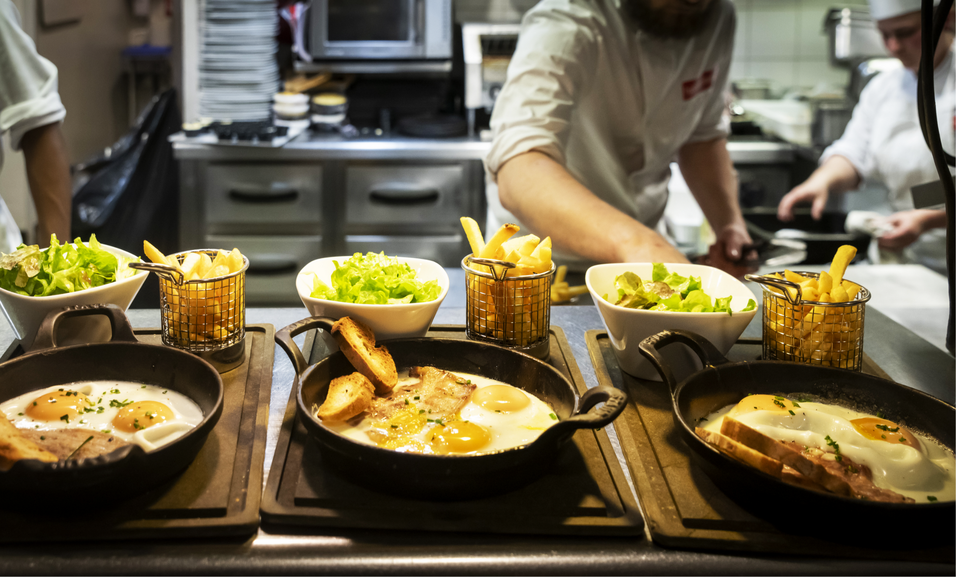 Cast iron pans of food, and baskets of fries sit on a counter in front of chefs.
