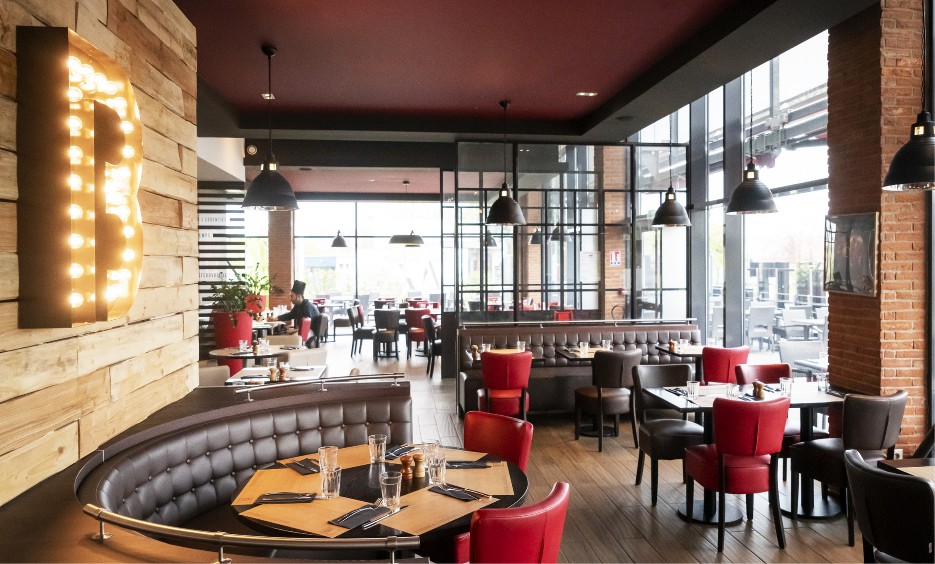 Restaurant interior with tables and chairs, booth seating, large windows, exposed brick, and a lit-up letter