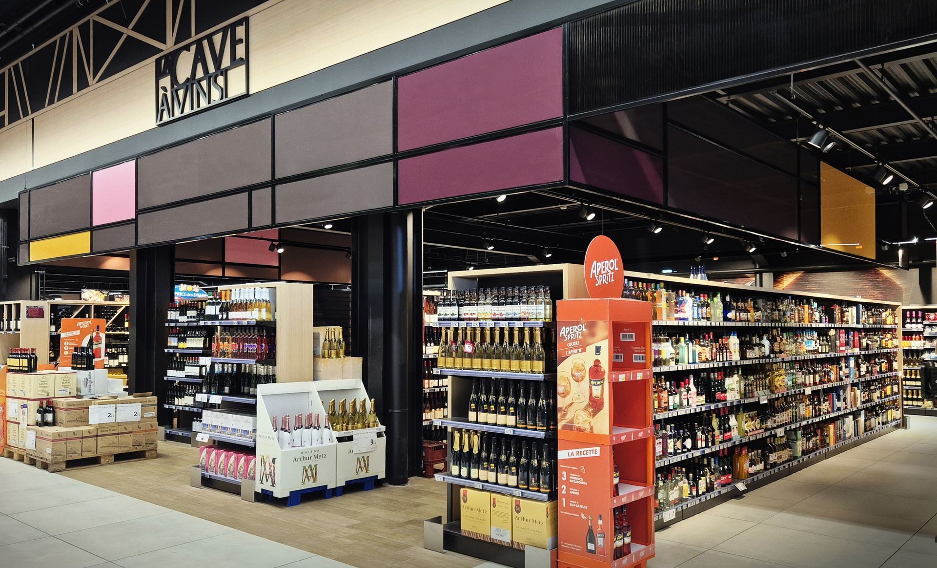 Entrée rayon vins avec des étagères de produits et un présentoir rouge sous un auvent noir et rose.