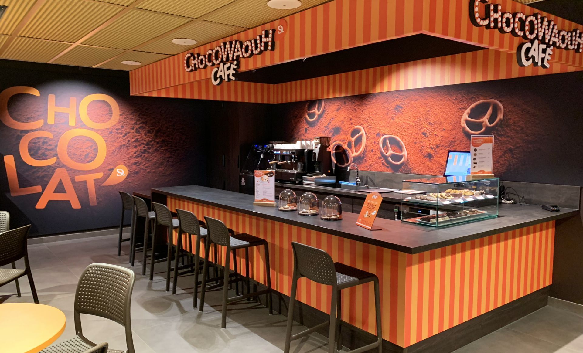 Chocolate cafe interior with counter seating. Orange and black striped decor, coffee machine, baked goods, dark walls.