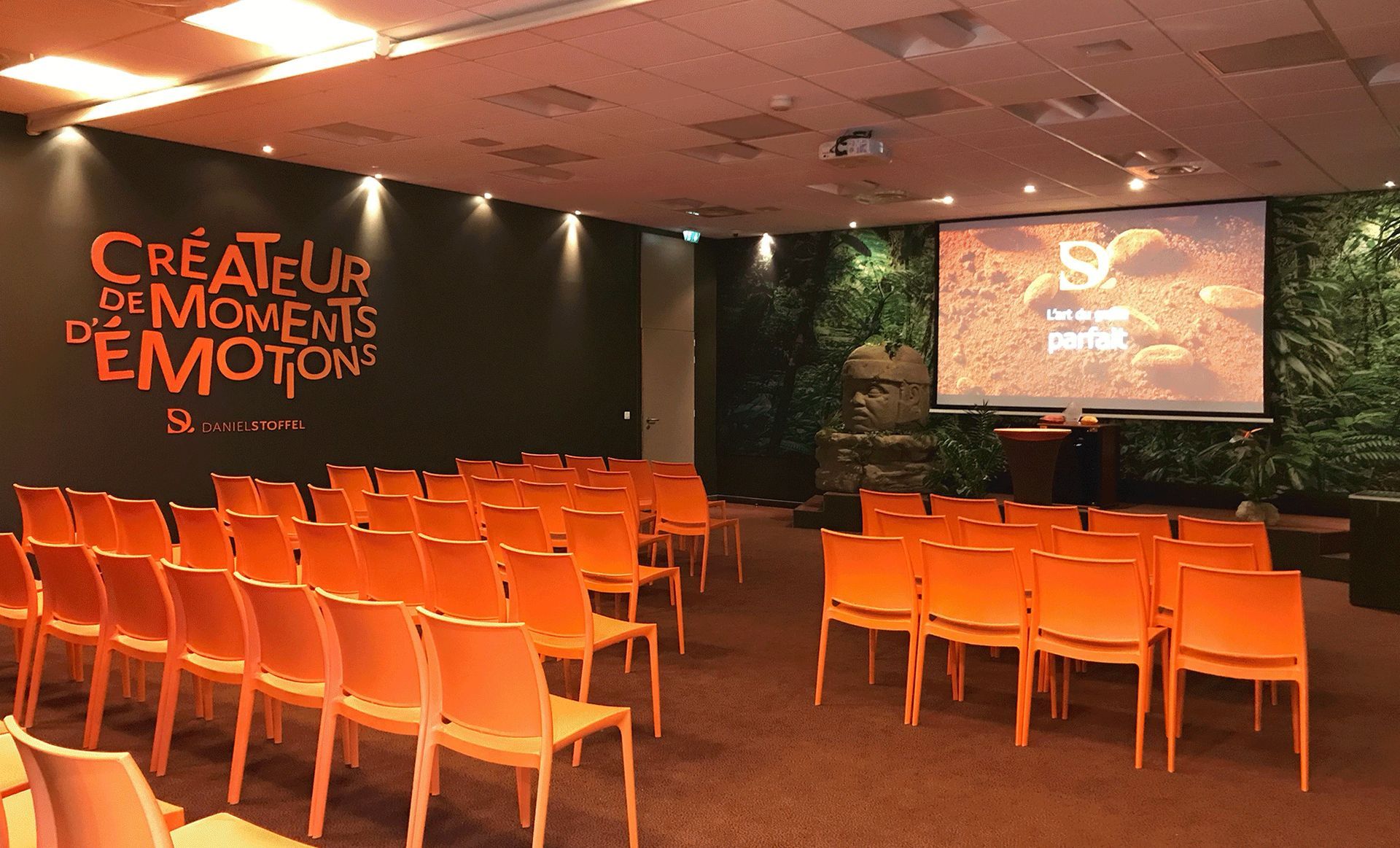 Orange chairs face a projector screen in a conference room with a botanical wall.