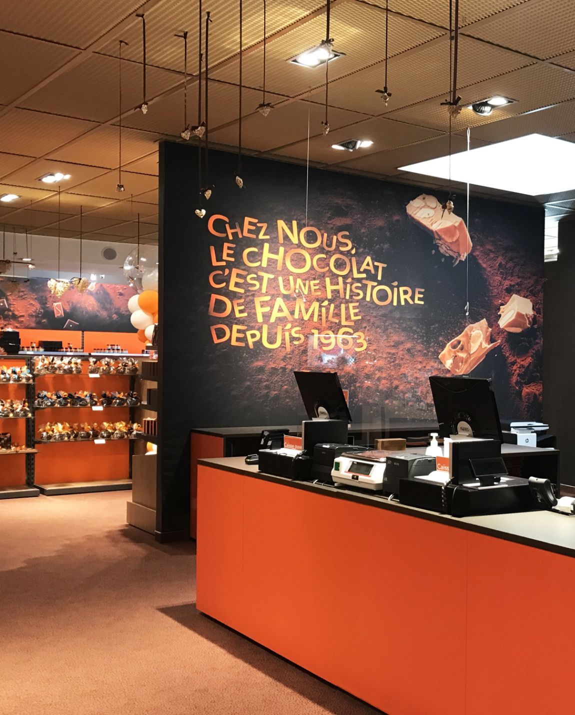 Interior of a chocolate shop with an orange and dark brown color scheme, a cash register, and a sign.