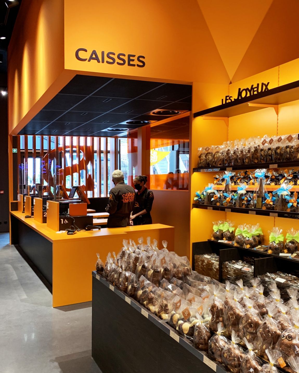 Interior of a chocolate shop with orange accents. Counters, shelves with packaged treats, and staff behind the counter.