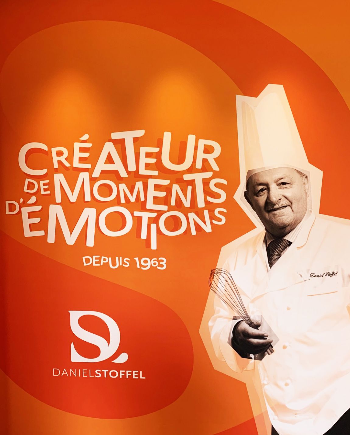 Chef holding a whisk, smiling, and wearing a tall hat and white coat. Orange background with text and logo.