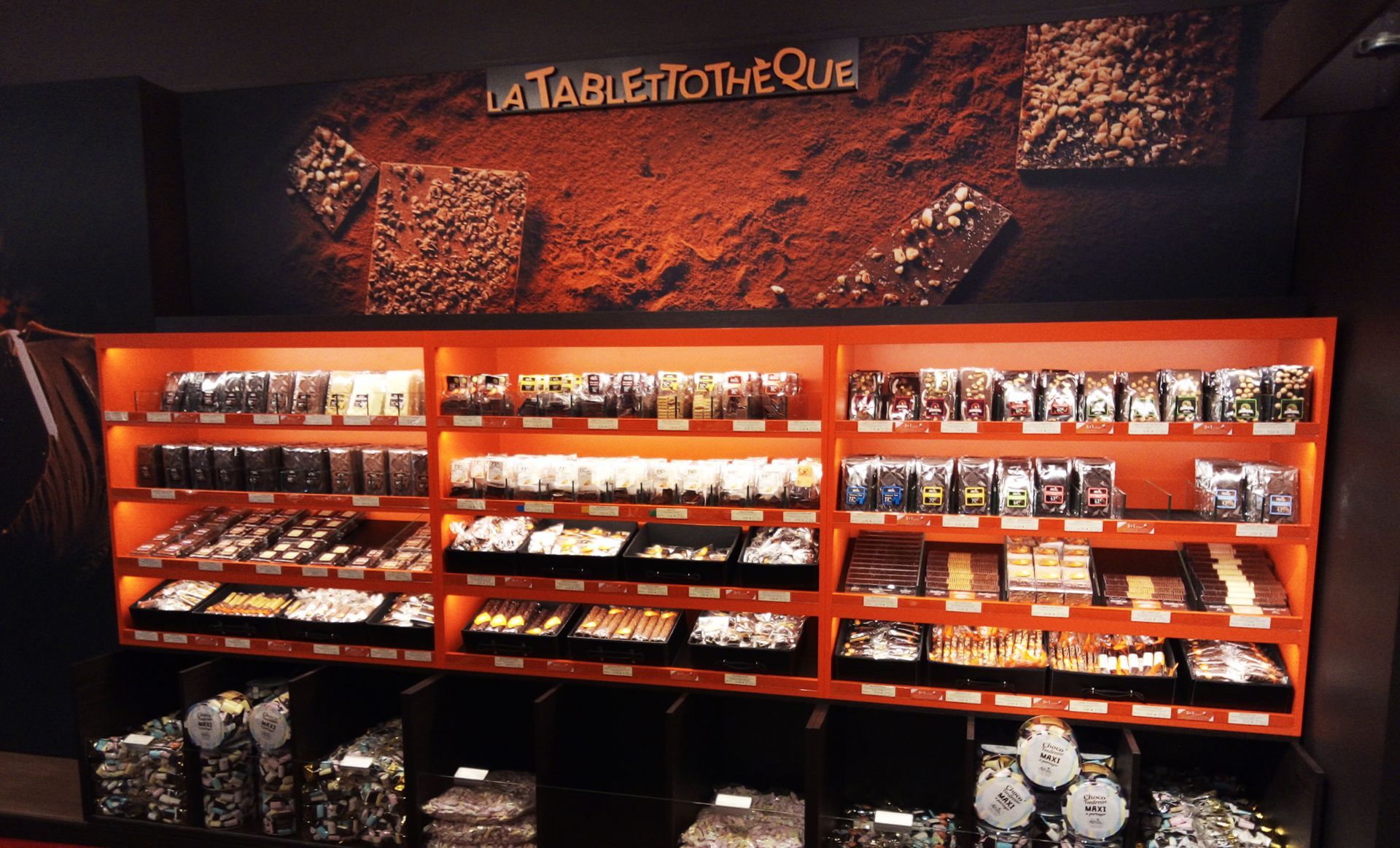 Shelves of chocolate bars and treats in an orange display case. Brown backdrop with chocolate pieces.