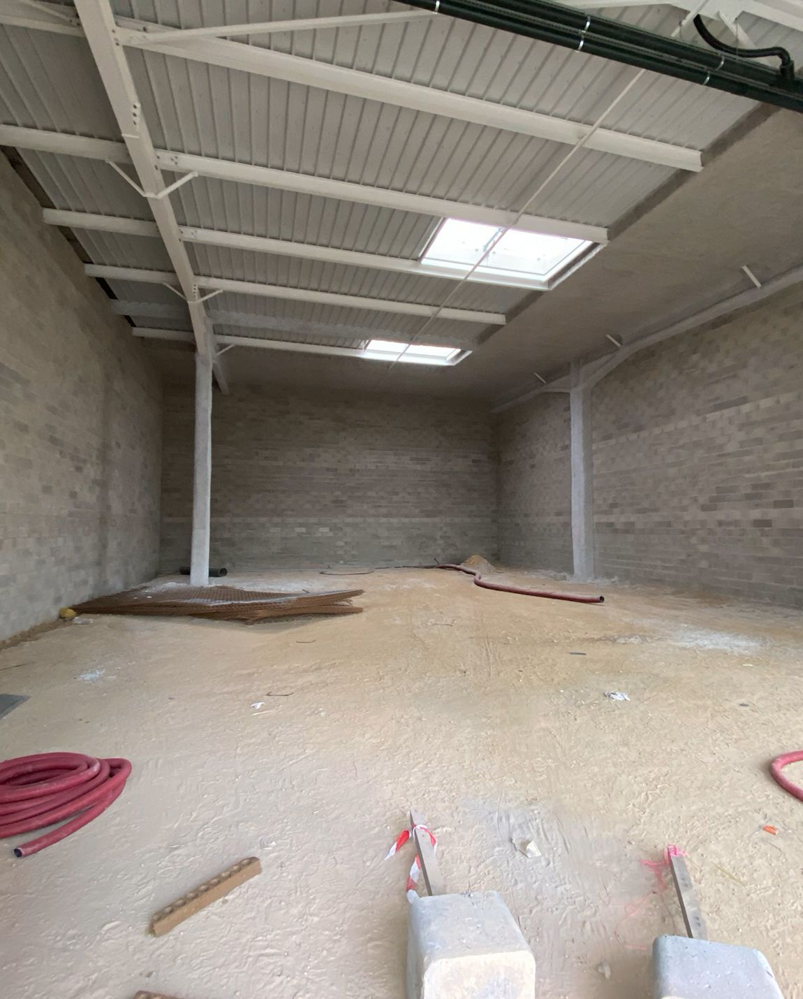 Empty warehouse interior with concrete walls, metal roof, and skylights. Construction materials visible on the floor.