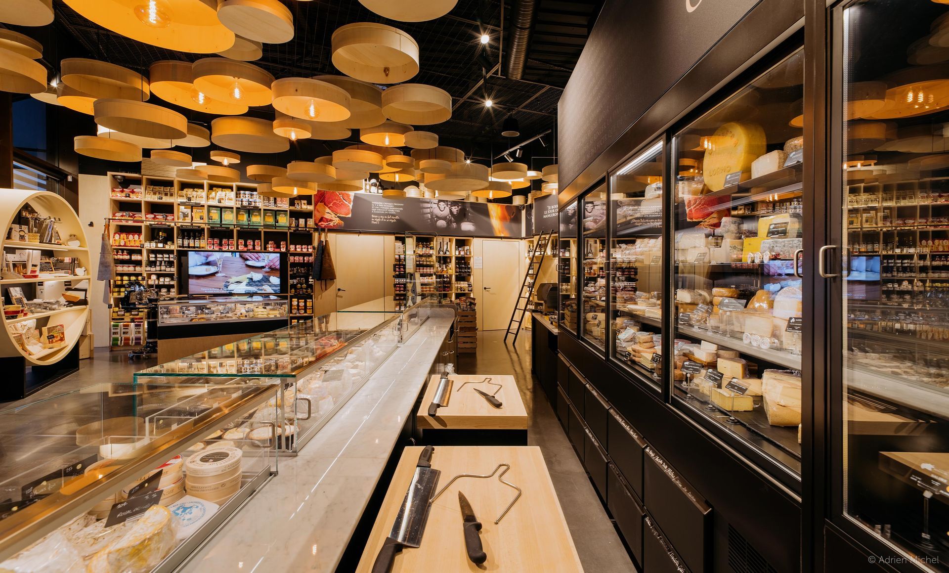 Inside a cheese shop: refrigerated cases, long display counter, hanging decorations, various food items.