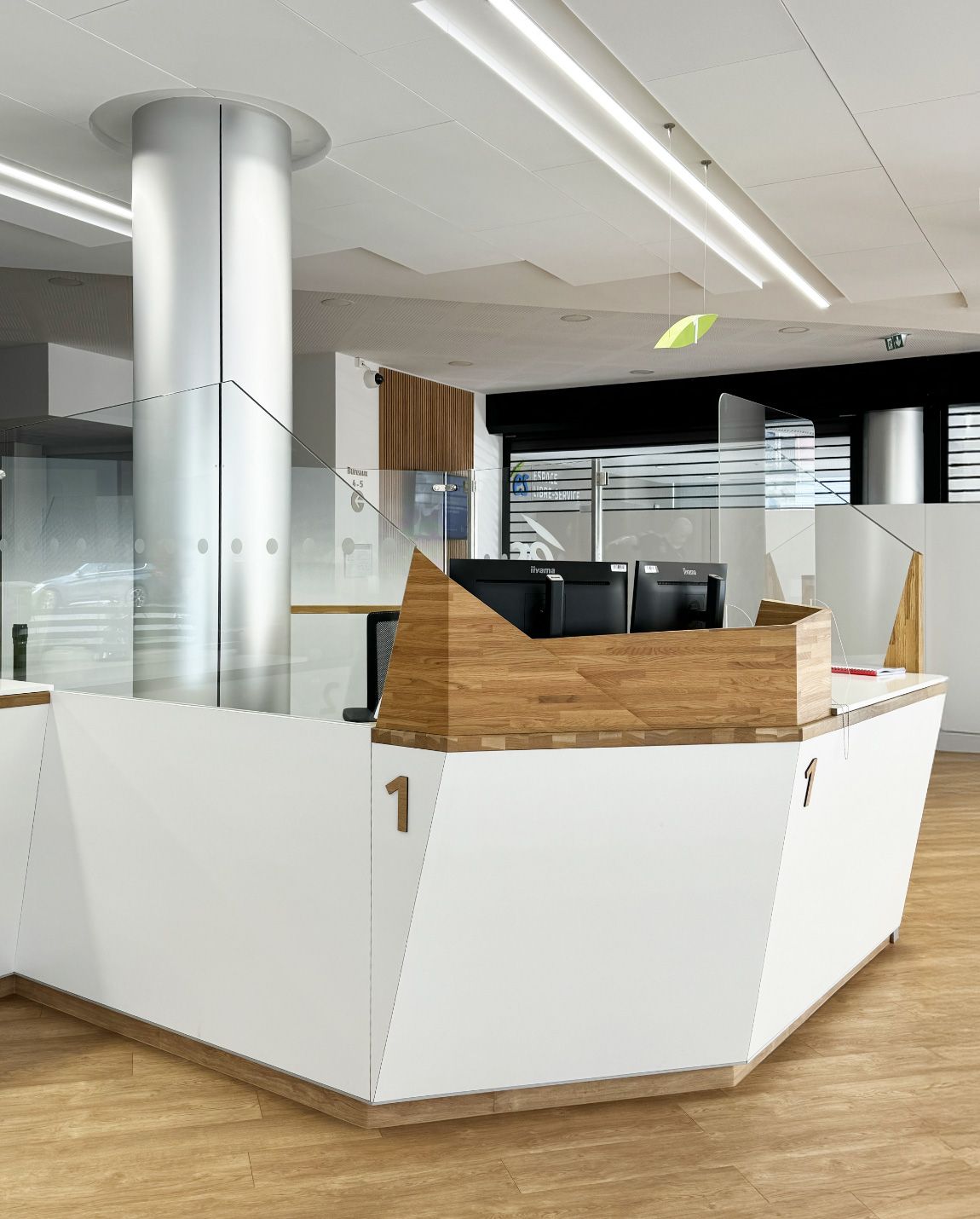 Modern white and wood reception desk in an office. A pillar and glass dividers are visible. Number 1 is on the desk.